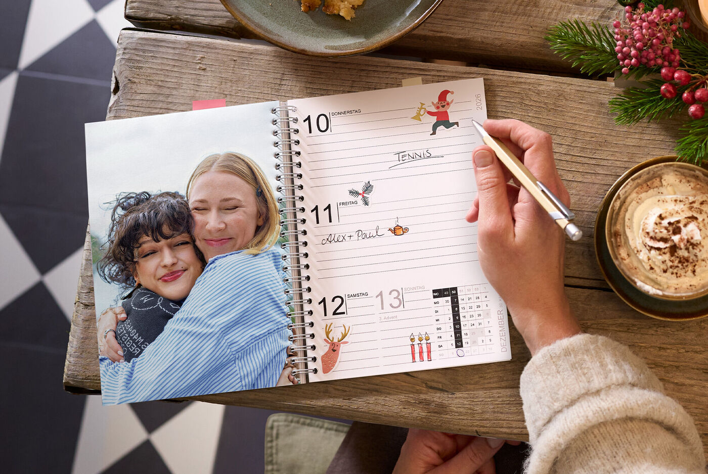 Offener CEWE Fotokalender mit Terminseite und Foto zweier Freundinnen auf einem Holztisch neben Tasse und Tannenzweig