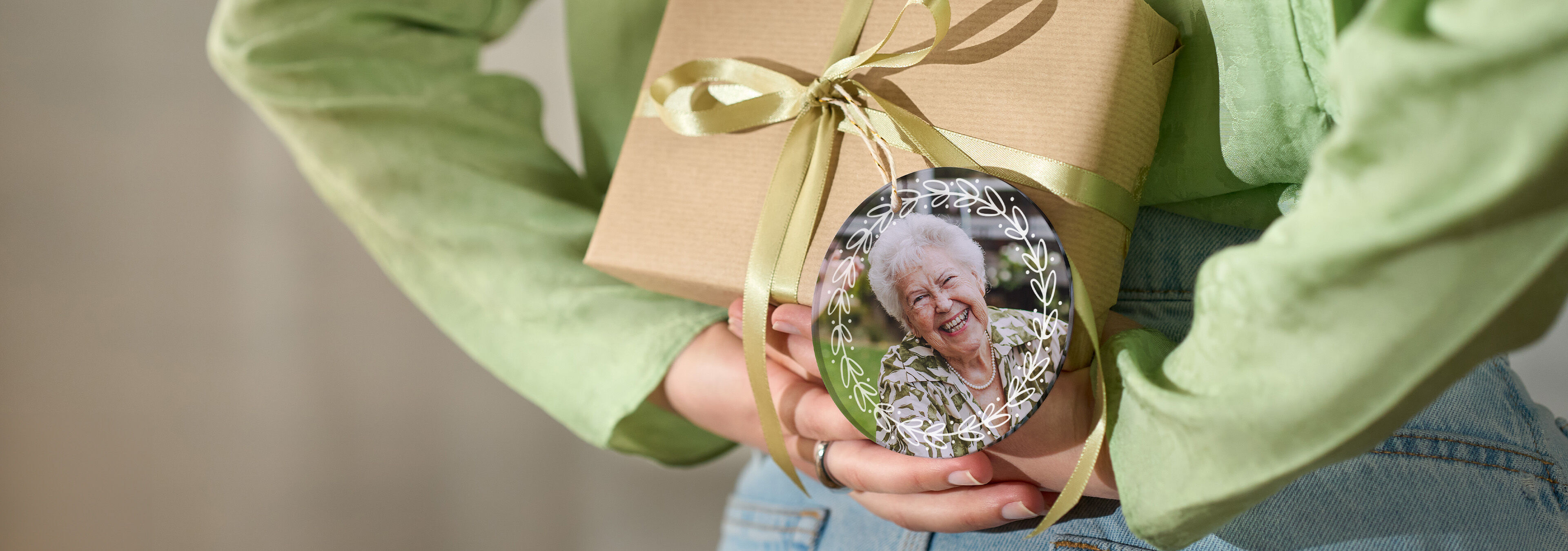 Person hält ein Geschenk mit Schleife und rundem Anhänger mit Foto einer lächelnden älteren Person hinter dem Rücken.