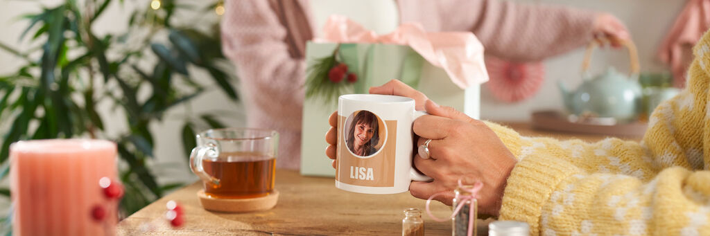 Person hält Tasse mit Foto und dem Namen Lisa neben einer Geschenkbox.
