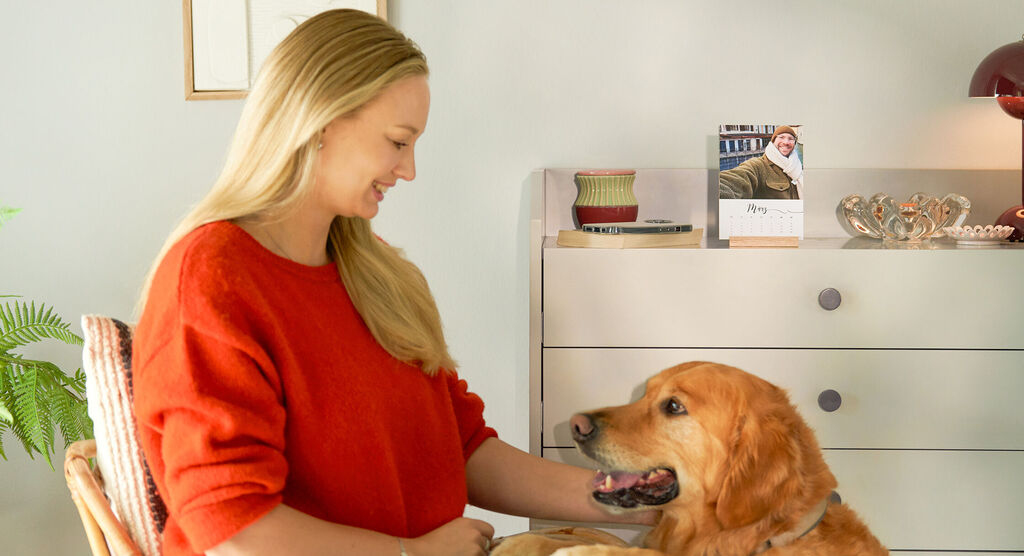 Person sitzt mit einem Hund im Wohnzimmer. Auf der Kommode steht ein CEWE Fotokalender