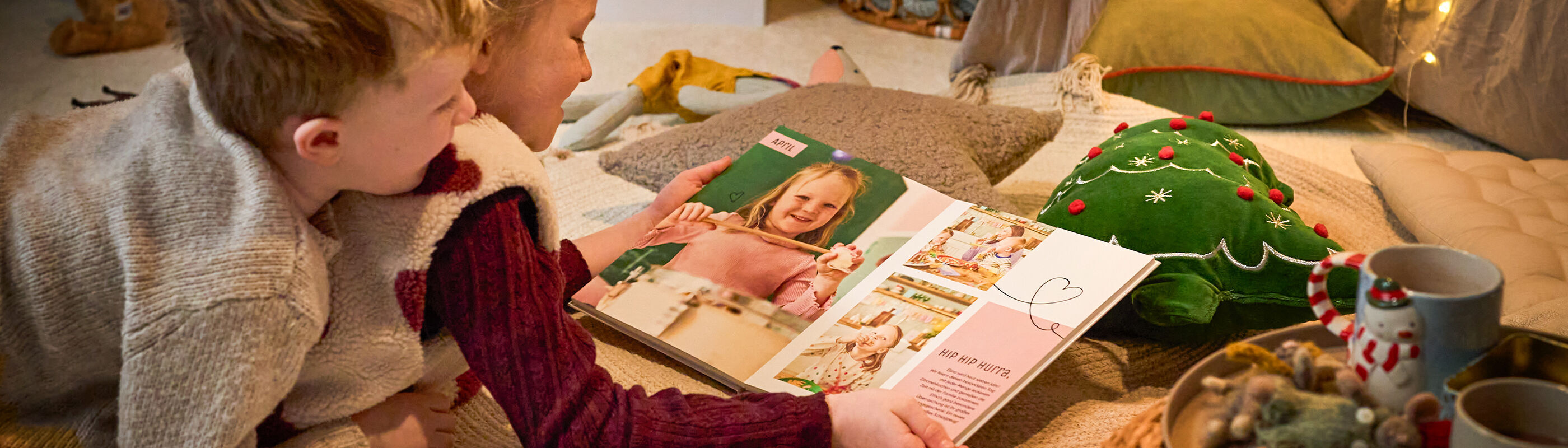 Zwei Kinder betrachten gemeinsam ein CEWE FOTOBUCH mit Familienfotos in gemütlicher, weihnachtlicher Umgebung.