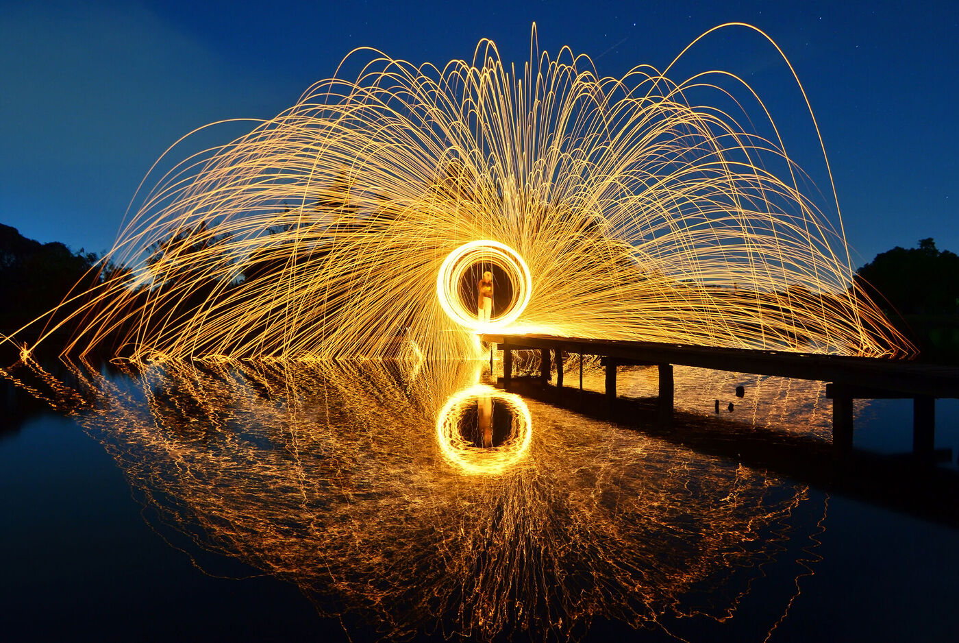 Eine Person steht auf einem Holzsteg und erzeugt mit Feuerwerk funkelnde Lichtspuren über dem Wasser in der Nacht. Die Lichtspuren reflektieren sich im Wasser und schaffen eine magische Atmosphäre.
