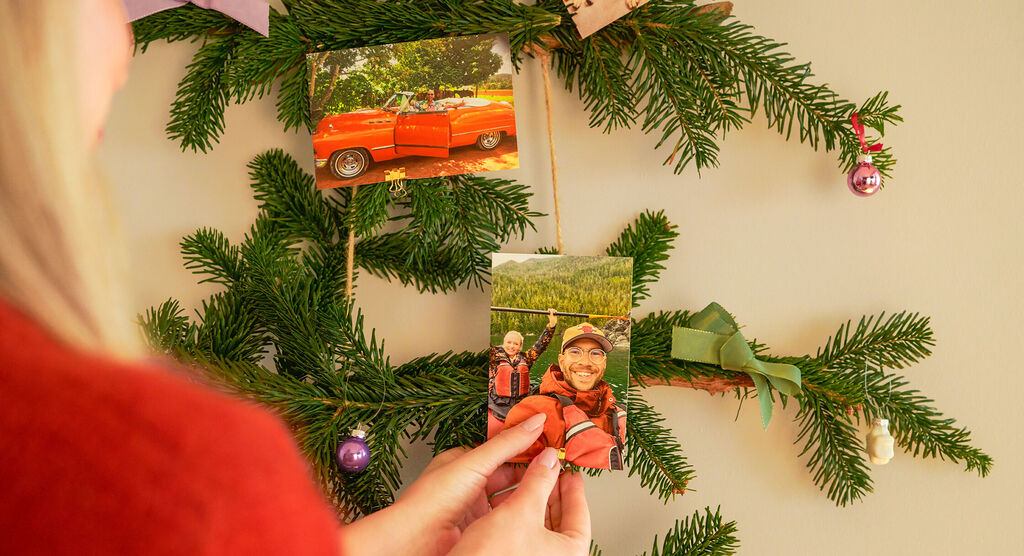 Person befestigt fröhlich Fotos an einem Tannenbaum aus Zweigen mit bunten Bändern und Kugeln