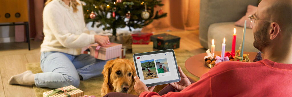 Frau packt Geschenke am Weihnachtsbaum ein, Mann sitzt mit Tablet und Hund daneben.