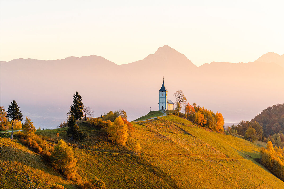 Schöne Landschaft mit einer kleinen Kirche auf einem Hügel, umgeben von bunten Bäumen und Bergen im Hintergrund
