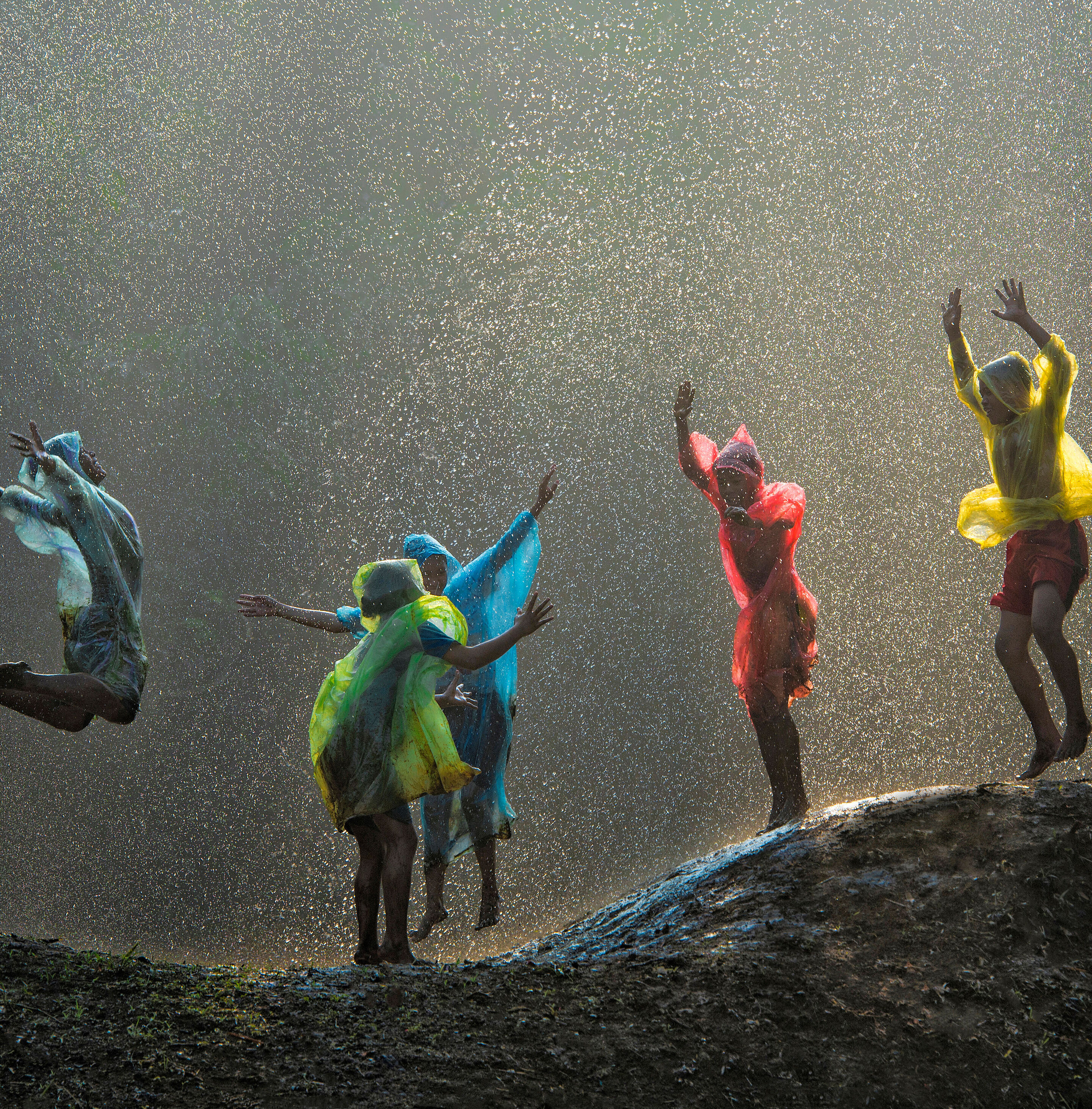 Fünf Kinder mit bunten Ponchos hüpfen im Regen auf einem Hügel.
