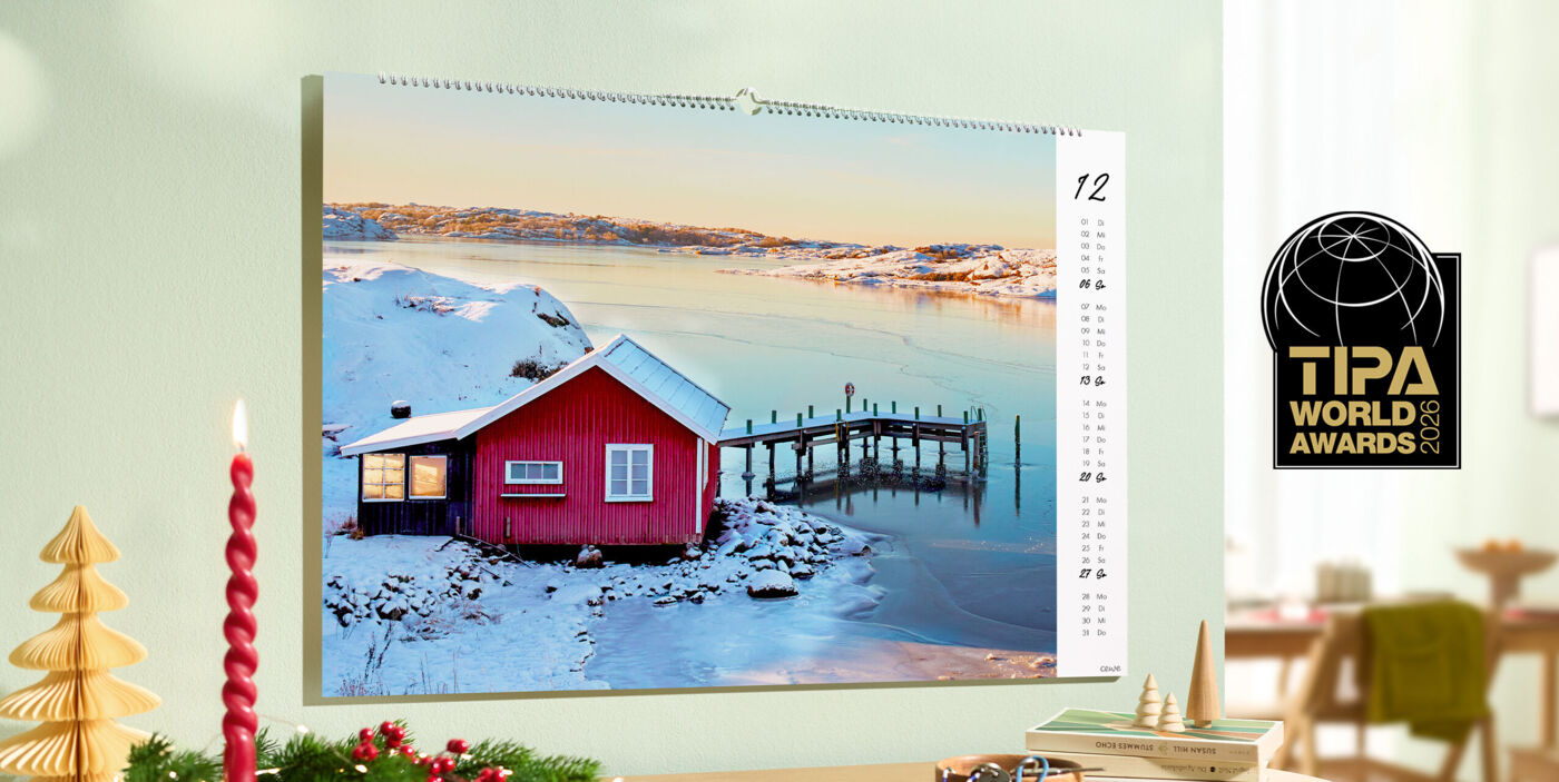 CEWE Kalender mit dem Bild eines roten Hauses an einem verschneiten See hängt an einer Wand in weihnachtlicher Umgebung, TIPA World Awards