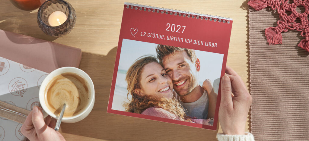 Kalender mit dem Titel ‚12 Gründe, warum ich dich liebe‘ auf einem Tisch, daneben eine Tasse Kaffee und Kerzenlicht
