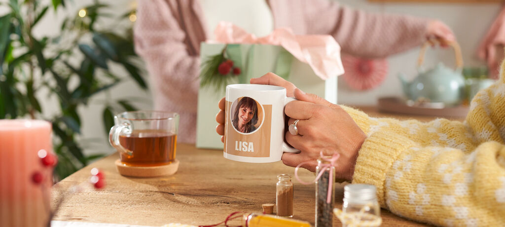 Person hält Tasse mit Foto und dem Namen Lisa neben einer Geschenkbox.