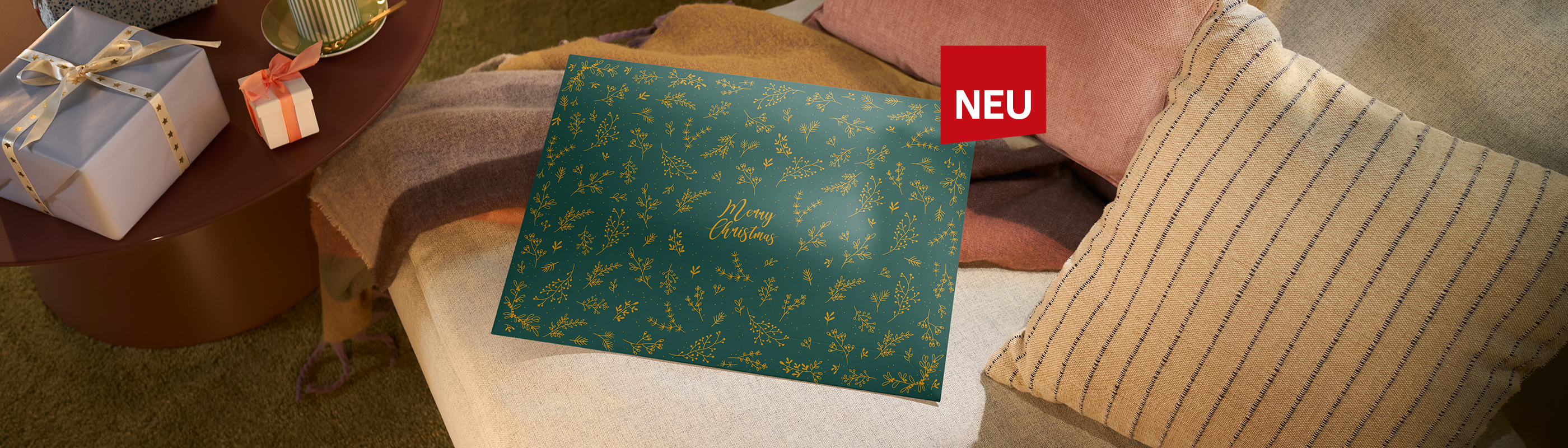 Grüne Geschenkverpackung für CEWE Fotokalender mit goldenen Zweigen und dem Schriftzug Merry Christmas, auf einem Sofa platziert.