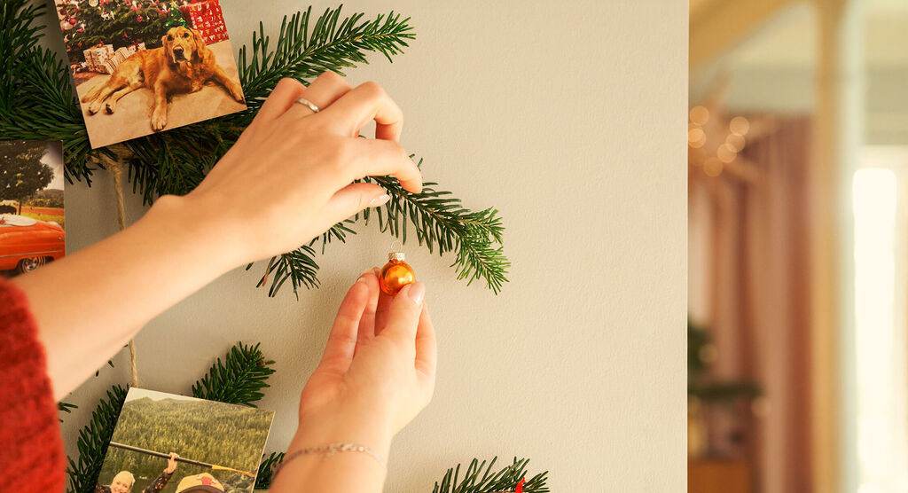 Zwei Hände schmücken einen Tannenast an der Wand mit einer kleinen orangefarbenen Kugel und Fotos