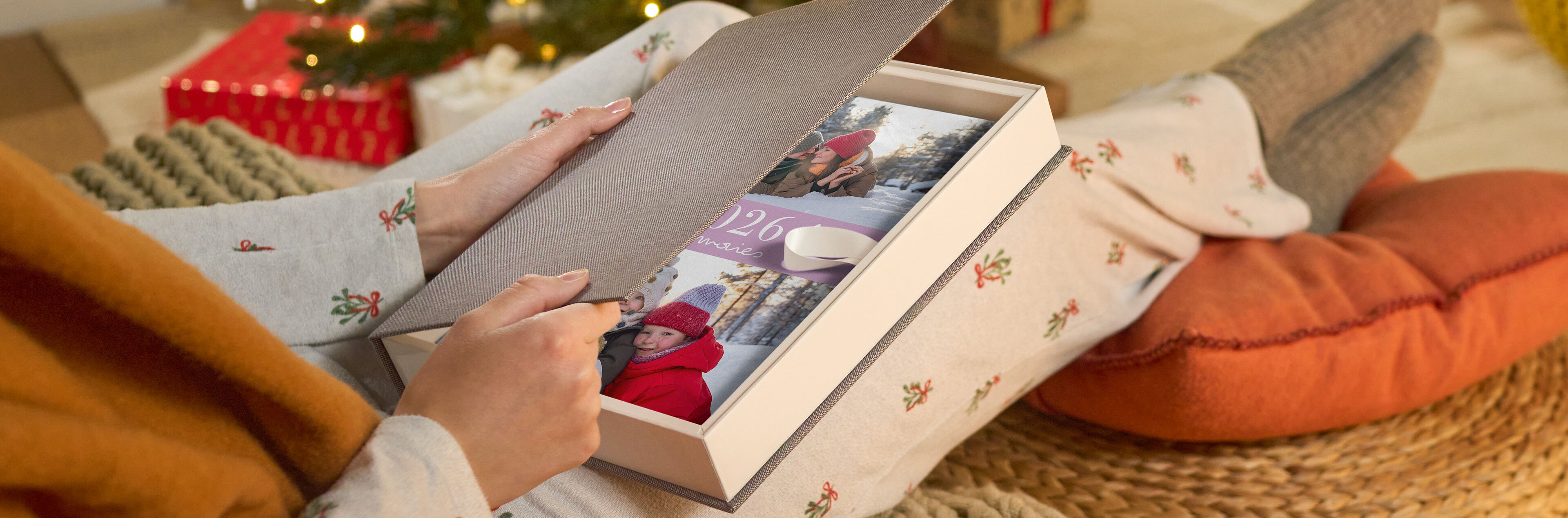 Eine Person öffnet eine Geschenkbox mit einem CEWE FOTOBUCH mit Winterfotos in weihnachtlicher Umgebung.
