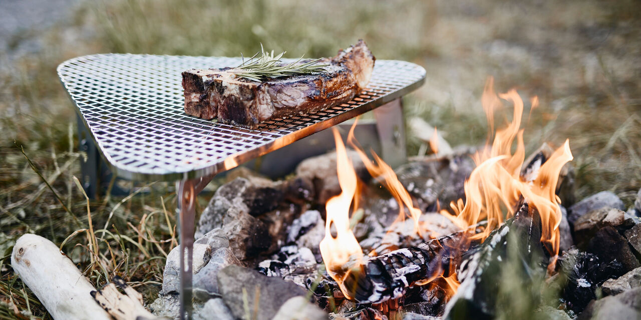 Ein saftiges Stück Fleisch grillt über einem offenen Feuer. Der Grill ist auf einem Steinhaufen platziert, umgeben von grüner Wiese.