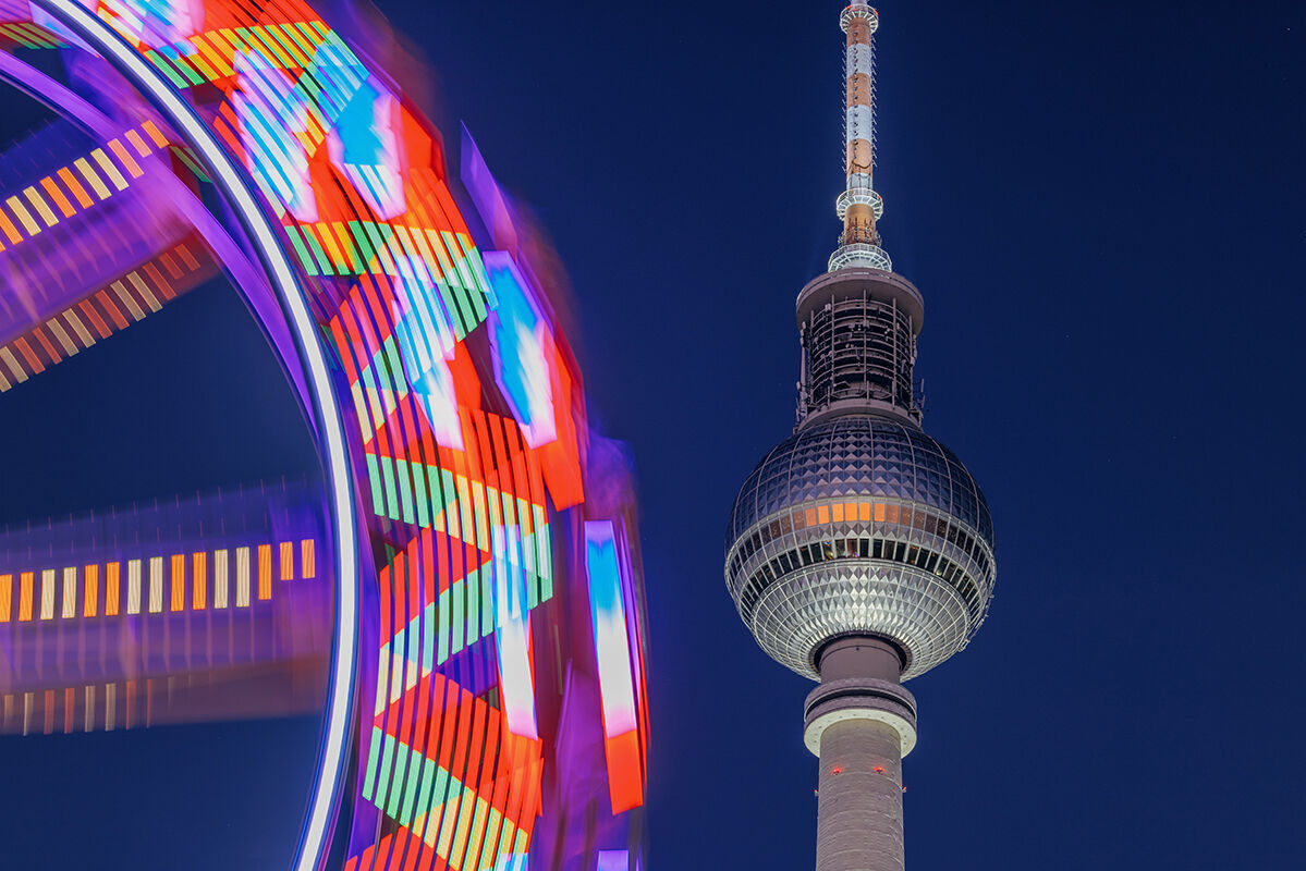 Ein farbenfrohes Riesenrad dreht sich im Vordergrund, während der Berliner Fernsehturm strahlend im Hintergrund leuchtet.