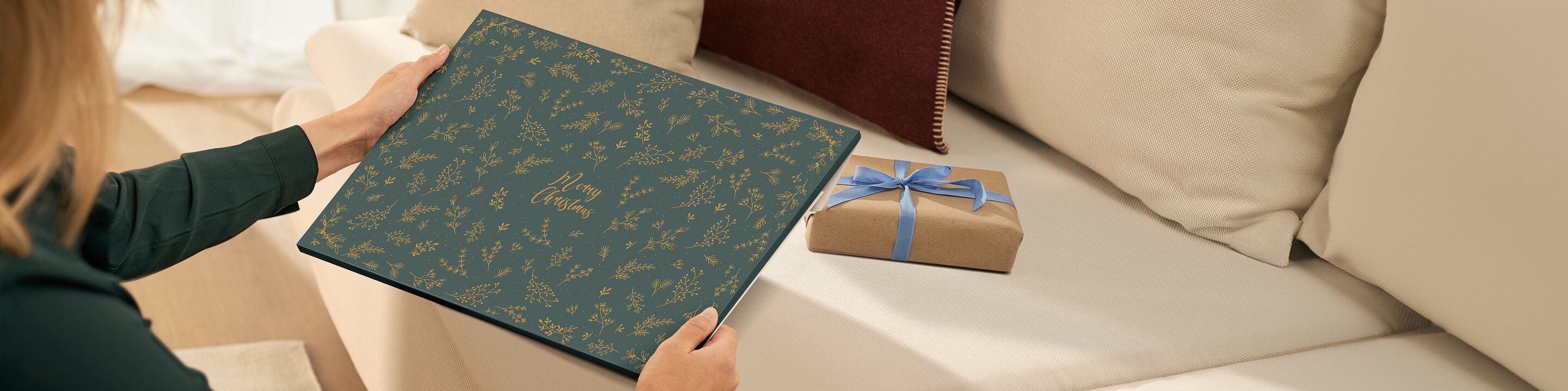 Eine Person hält ein CEWE Fotobuch mit grünem Cover auf dem Sofa, daneben liegt ein dekoriertes Geschenk.