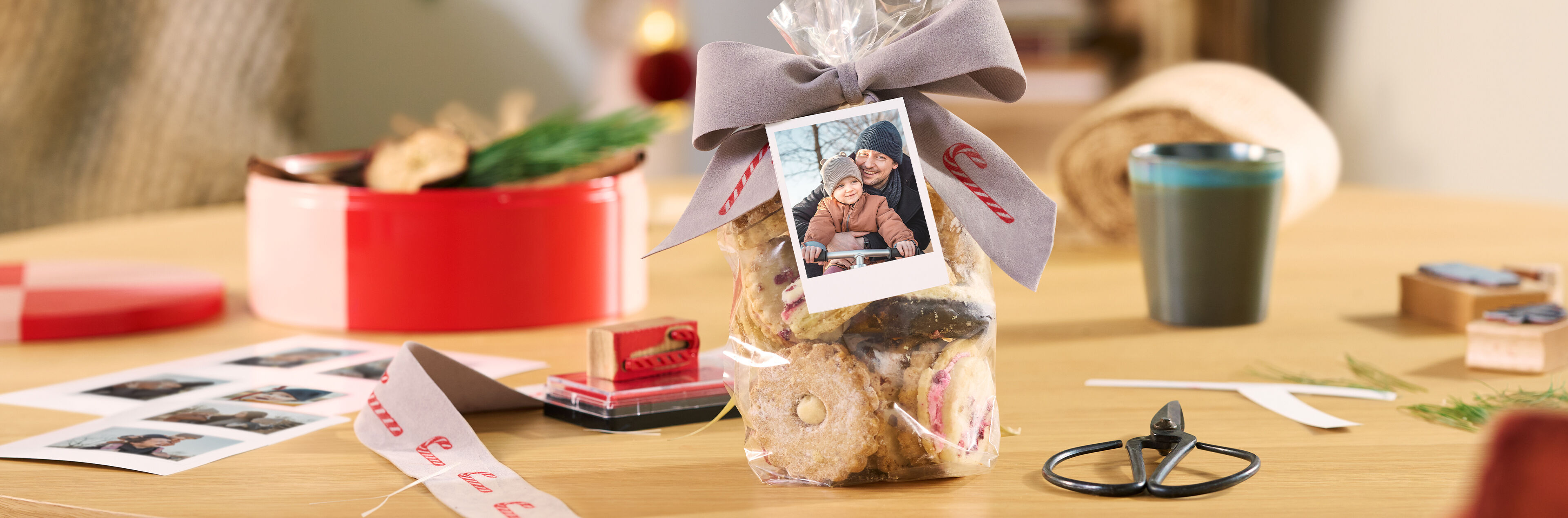 Geschenktüte mit Keksen, Schleife und Fotokarte auf einem Tisch, umgeben von Bastelzubehör.