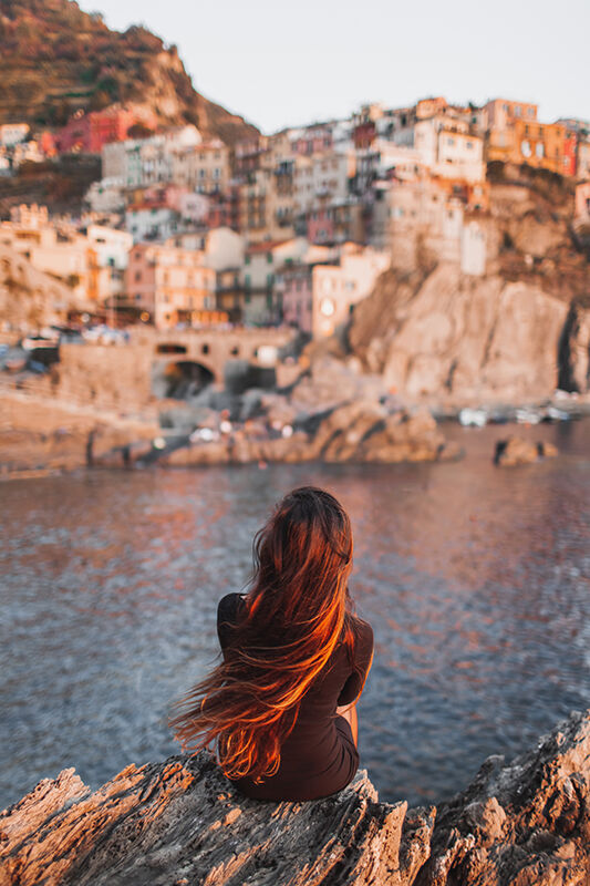 Frau mit langen Haaren sitzt am Felsen und schaut auf das Wasser und die Küstenstadt