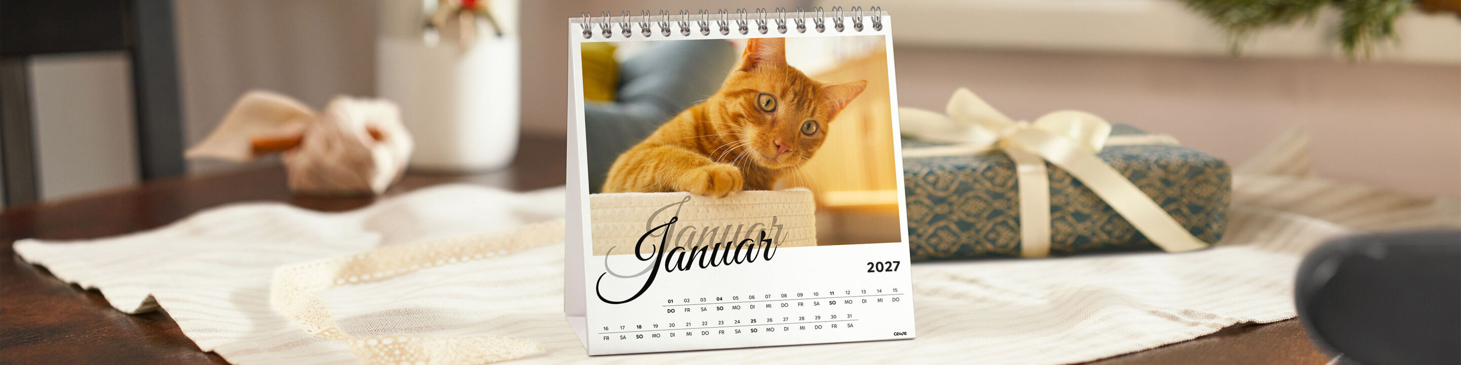 CEWE Tischkalender mit einem Foto einer orangefarbenen Katze, daneben ein Geschenk mit Schleife auf einem Holztisch