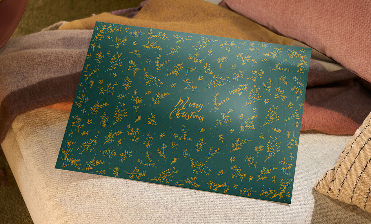 Grüne Geschenkverpackung für CEWE Fotokalender mit goldenen Zweigen und dem Schriftzug Merry Christmas, auf einem Sofa platziert.