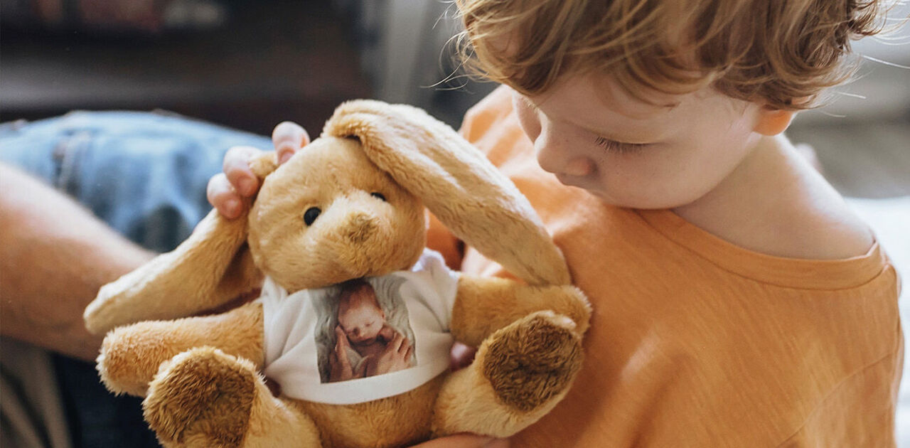 Kind hält einen Kuschelhasen mit Babyfoto auf dem T-Shirt in den Armen.