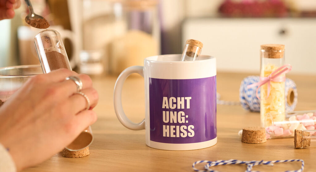 Tasse mit dem Text Achtung Heiss steht auf Holztisch zwischen Kakao und Zutaten.
