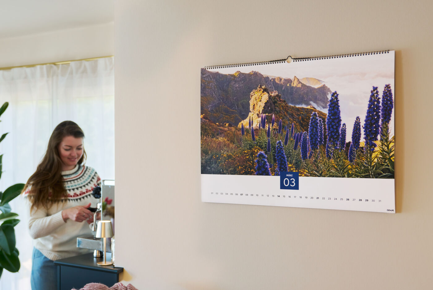 In einem Wohnzimmer hängt ein großer Wandkalender, im Hintergrund steht eine Frau an einer Kaffeemaschine und bereitet etwas zu.