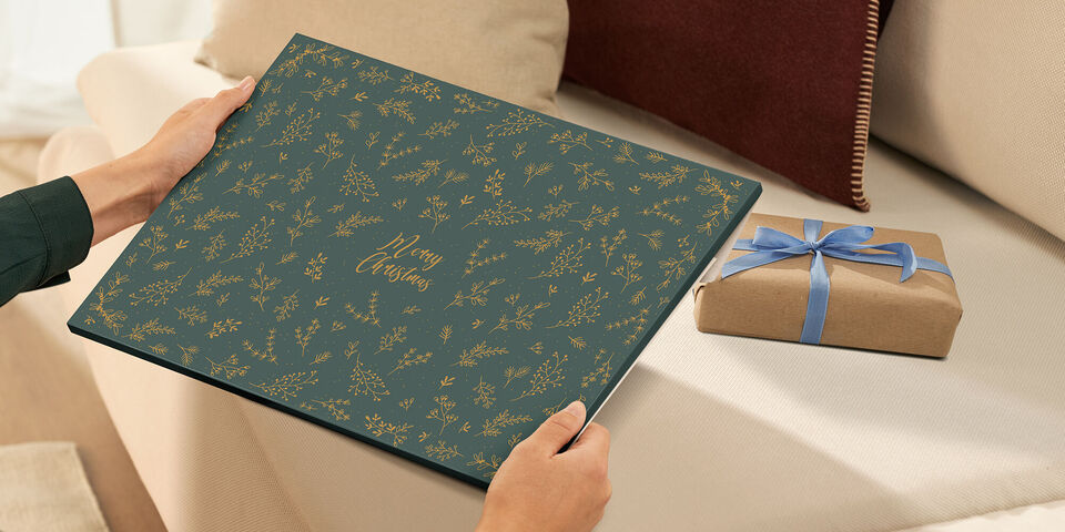 Eine Person hält ein CEWE Fotobuch mit einem schönen floralen Muster in den Händen. Neben ihr liegt ein liebevoll verpacktes Geschenk.