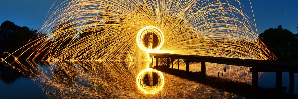 Ein Mensch steht auf einer Holzbrücke und zaubert mit brennenden Funken ein leuchtendes Muster in der Luft, während sich das Licht im Wasser reflektiert.