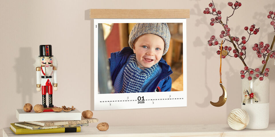 CEWE Fotokalender, aufgehängt an einer Holzleiste, neben Nussknacker-Deko, Walnüssen und einem Zweig mit roten Beeren