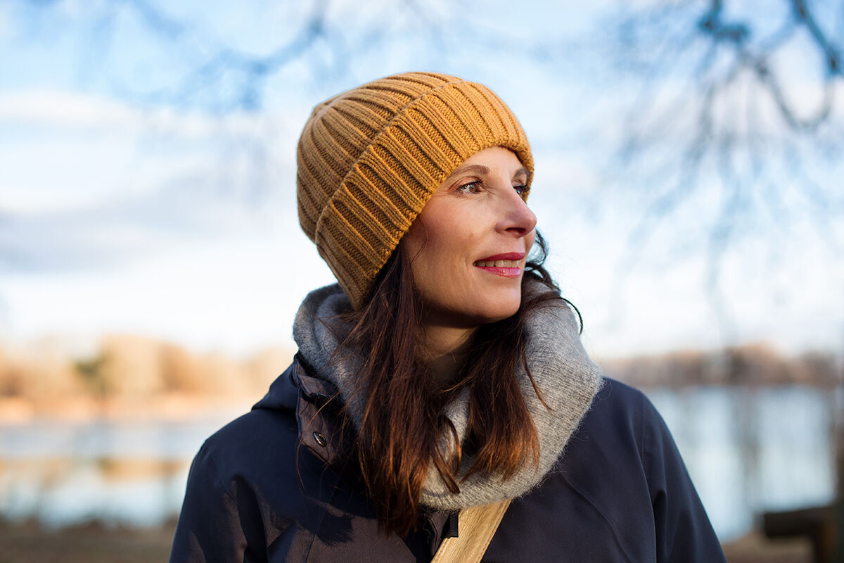 Eine Person mit Wollmütze und Jacke steht im Freien, im Hintergrund sind Bäume und Wasser zu sehen