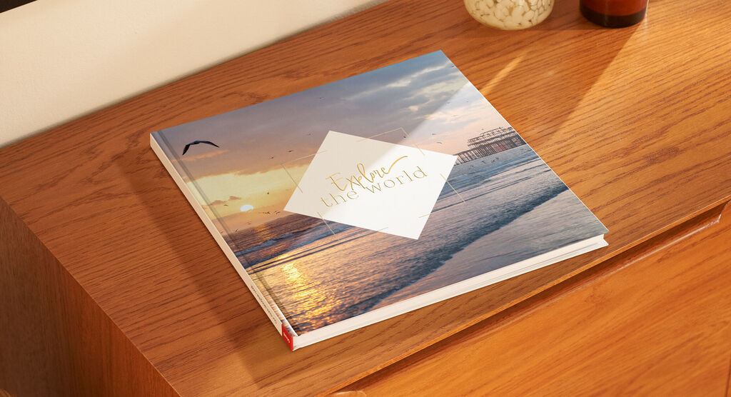 Ein CEWE FOTOBUCH liegt auf einem hölzernen Tisch. Das Cover zeigt eine Strandlandschaft mit Sonnenuntergang und dem Text 'Explore the World'.