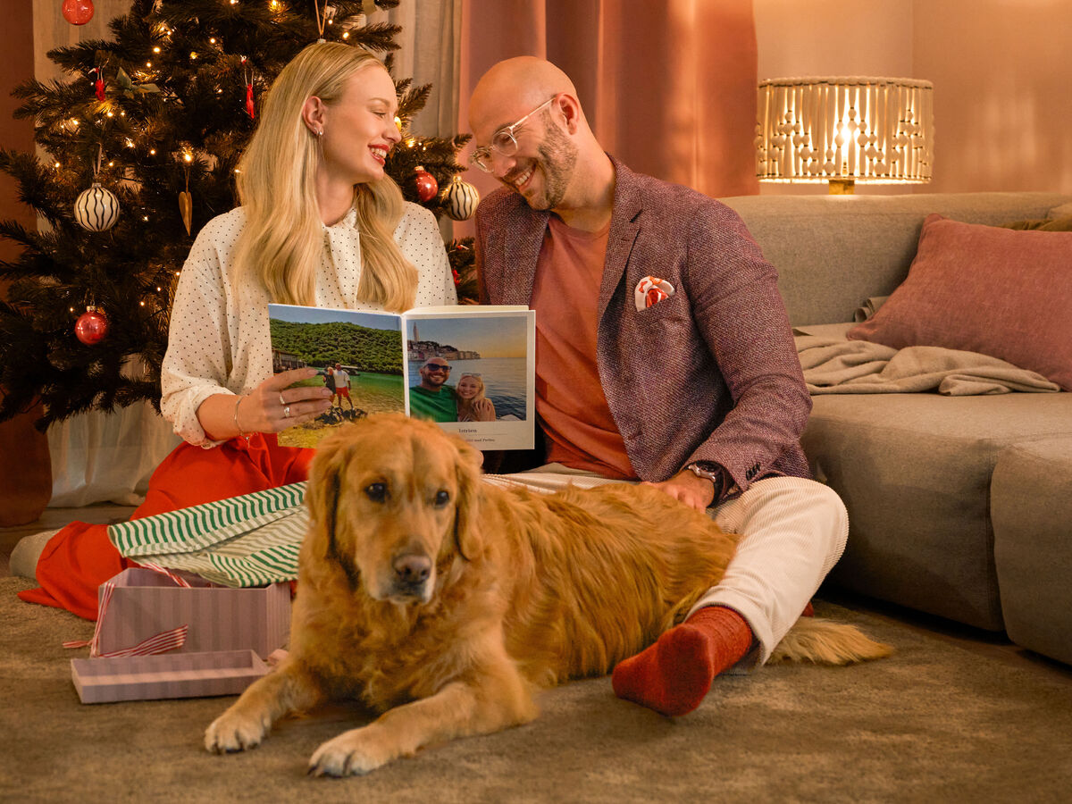 Zwei Menschen sitzen vor dem Weihnachtsbaum, halten ein CEWE FOTOBUCH in den Händen, daneben liegt ein Hund und Geschenkpapier.