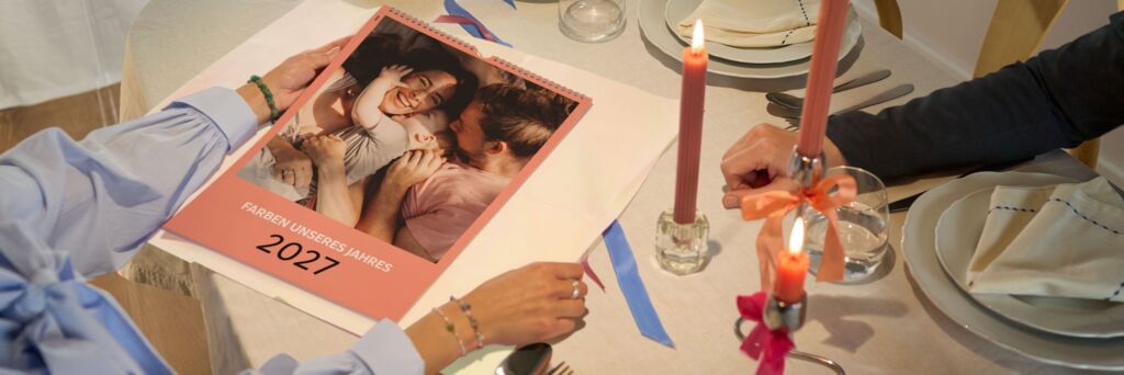 Zwei Personen am festlich gedeckten Tisch betrachten einen CEWE Fotokalender mit Familienfoto und Jahreszahl 2026