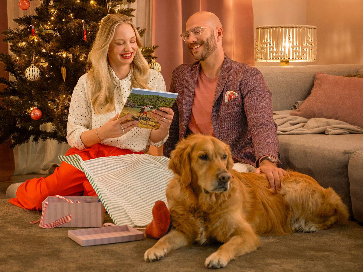 Zwei Menschen sitzen mit Hund vor dem geschmückten Weihnachtsbaum und freuen sich über ein Fotobuchgeschenk
