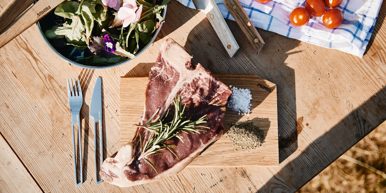 Großes Stück Fleisch auf einem Holzbrett mit Gewürzen und einer bunten Salatschüssel im Hintergrund.