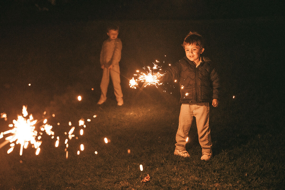 Zwei Kinder stehen in der Dunkelheit, eines hält ein Feuerwerk in der Hand und erfreut sich an den funkelnden Lichtern.