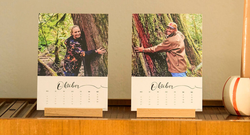 Zwei CEWE Fotokalender für Oktober. Je eine Person umarmt einen großen Baum im Wald
