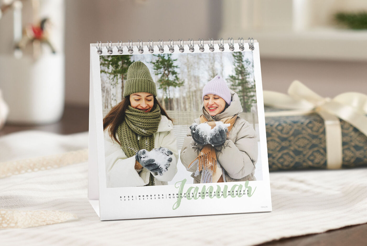 CEWE Tischkalender mit Januar-Motiv: Zwei Personen in winterlicher Kleidung, Schneeball in den Händen, daneben ein Geschenk