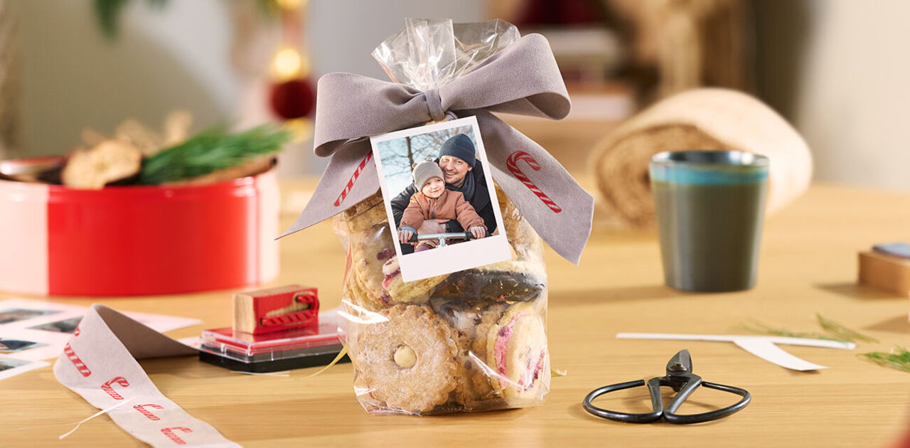 Geschenktüte mit Keksen, Schleife und Fotokarte auf einem Tisch, umgeben von Bastelzubehör.