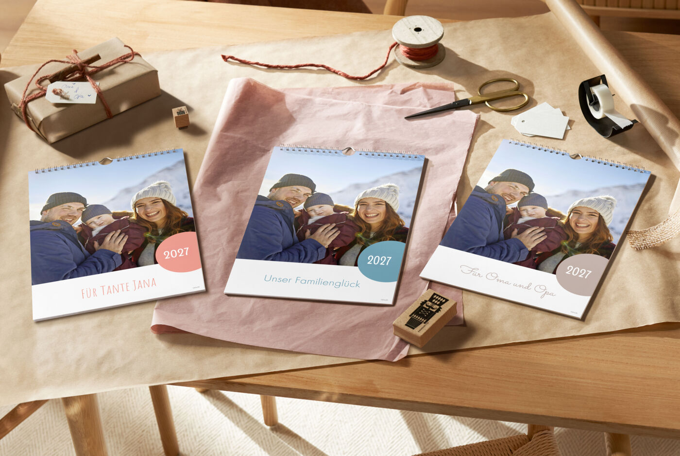 Drei individuelle CEWE Fotokalender liegen auf einem Tisch, verpackt in Geschenkpapier, mit einer Schere und Zubehör daneben.