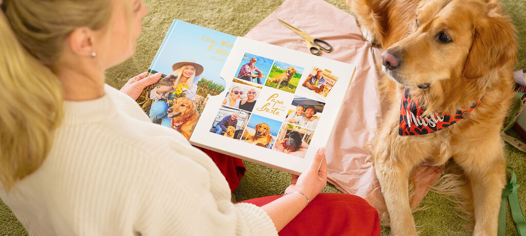 Eine Frau hält ein CEWE FOTOBUCH mit Familienfotos in den Händen. Vor ihr liegt ein Hund und schaut sie an.