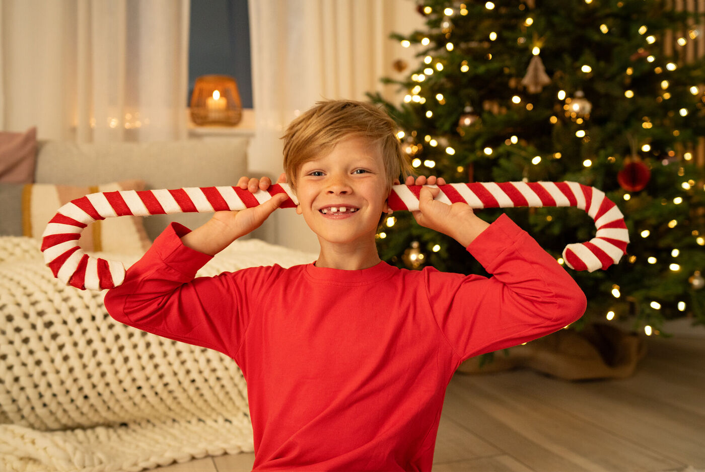 Ein Junge trägt einen langen, gestreiften Zuckerstangen-Requisit und blickt fröhlich umher. Im Hintergrund steht ein festlich geschmückter Weihnachtsbaum.