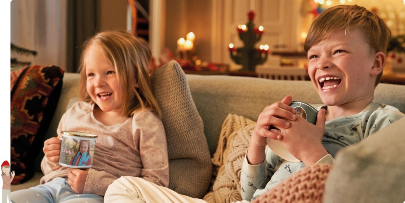Zwei lachende Kinder sitzen in weihnachtlicher Umgebung auf einem Sofa und halten jeweils eine Fototasse mit persönlichem Foto in der Hand.