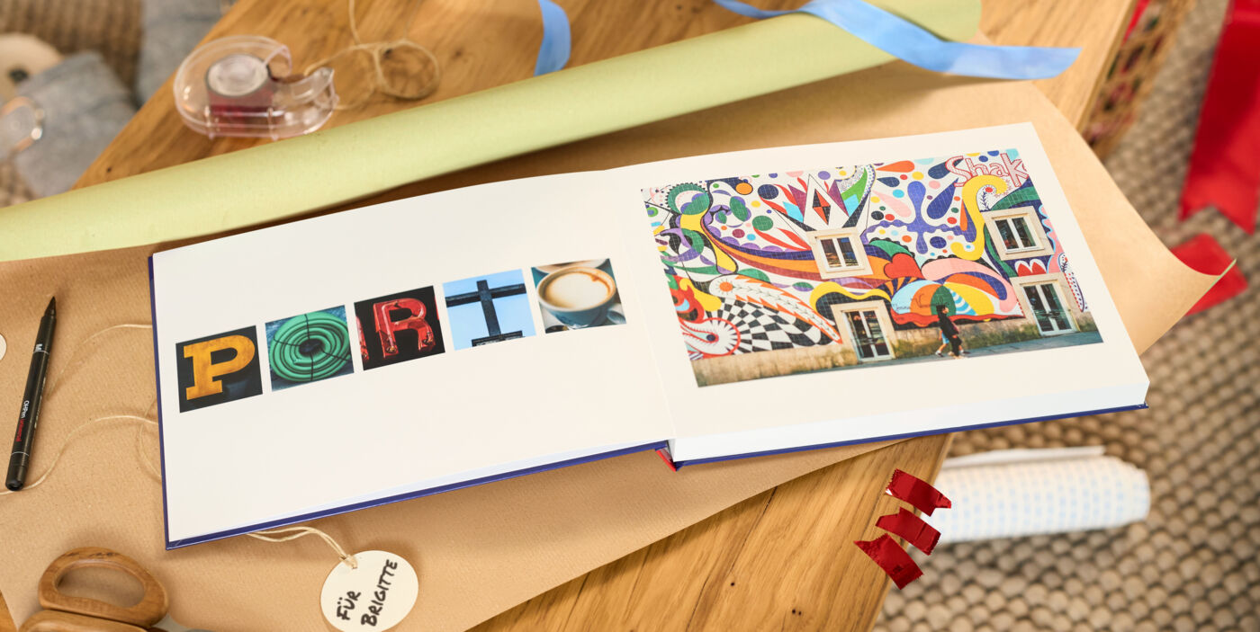 Offenes CEWE FOTOBUCH mit buntem Wandbild und Buchstaben-Collage auf Holztisch