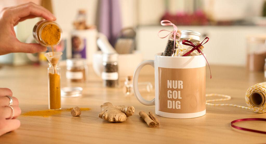 Hand füllt Kurkuma in Glasröhre, daneben Tasse mit Aufschrift NUR GOLDIG und Gewürzen.
