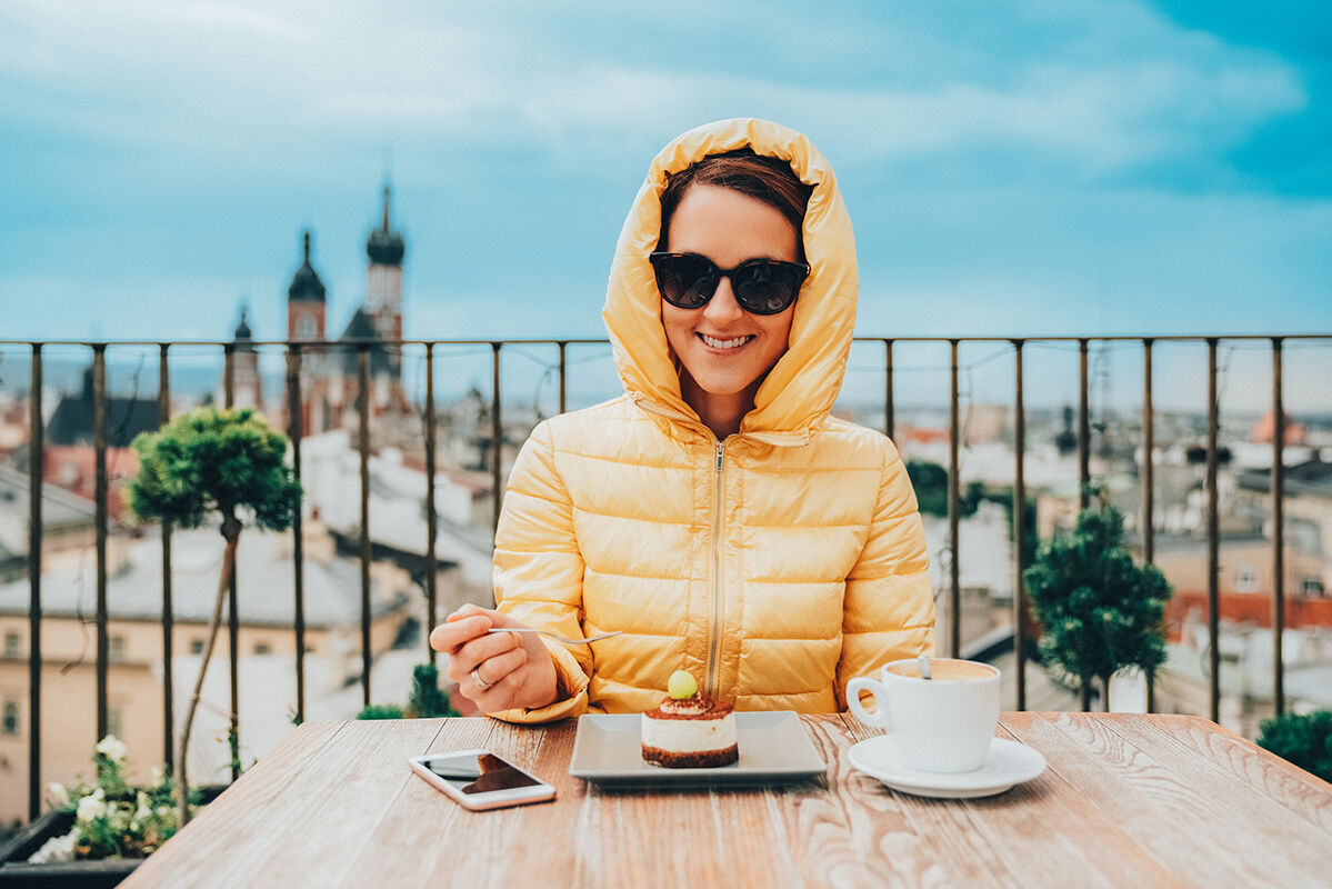 Junge Person in gelber Jacke genießt ein Dessert und eine Tasse Kaffee in einem Café mit schöner Aussicht auf die Stadt.