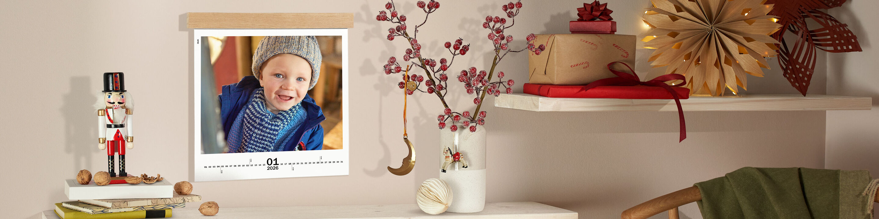 CEWE Fotokalender aufgehängt an einer Holzleiste, neben dekorativem Nussknacker, Walnüssen, roten Beerenzweigen und Geschenken