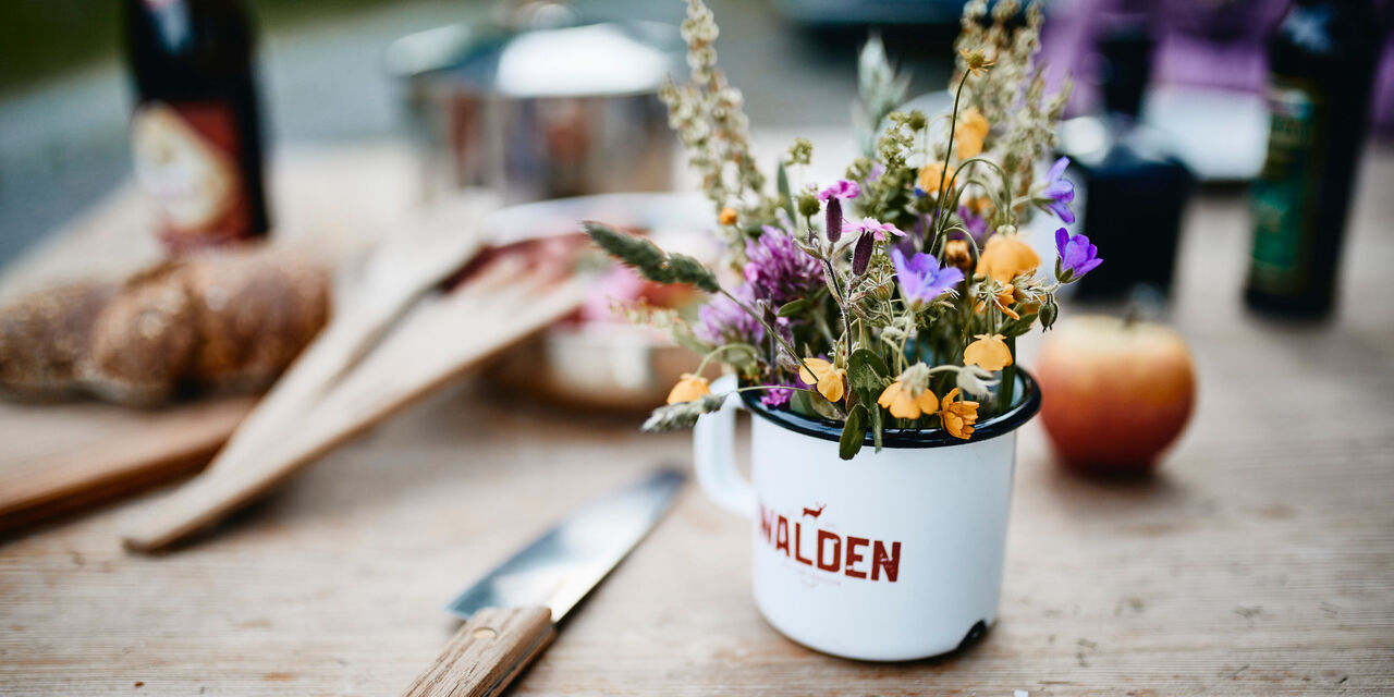 Ein bunter Strauß von Wildblumen in einem weißen Topf mit roter Aufschrift steht auf einem Holztisch. Im Hintergrund sind Küchenutensilien und eine Apfel sichtbar