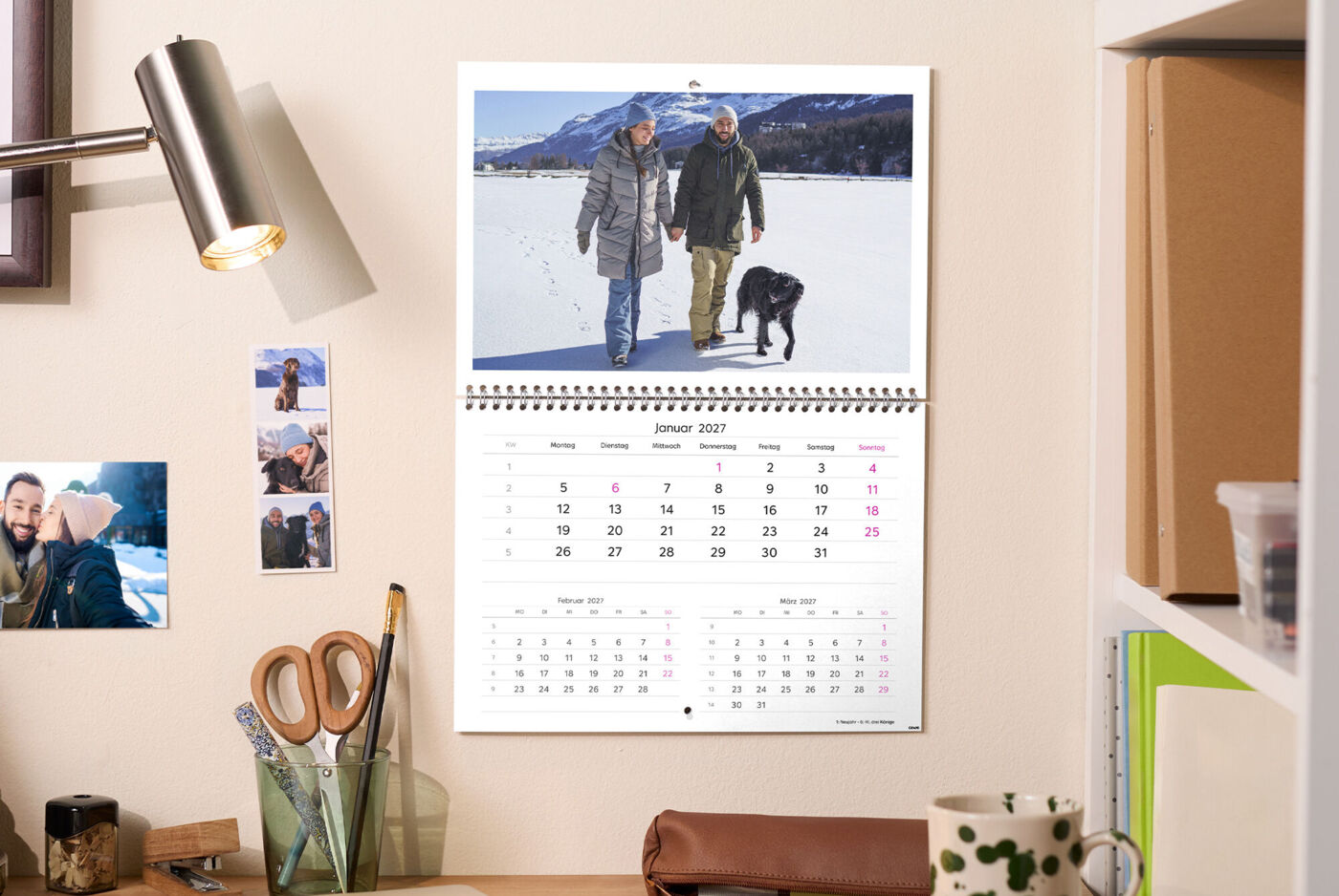 CEWE Fotokalender mit Paar und Hund beim Spaziergang im Schnee, Schreibtischlampe und Fotos daneben