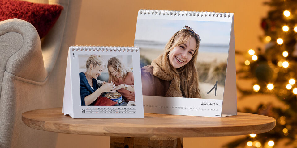 Zwei CEWE Fotokalender auf einem Holztisch, weihnachtliche Beleuchtung im Hintergrund